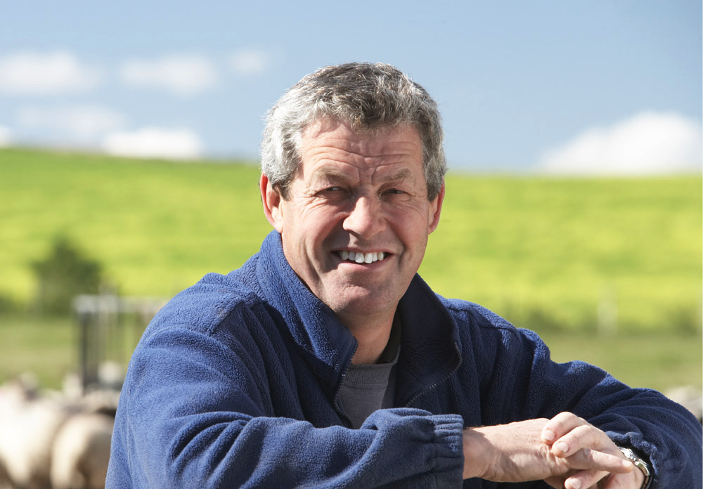 A person with short gray hair and a blue jacket smiles outdoors, standing in front of a blurred background of a green field and blue sky.